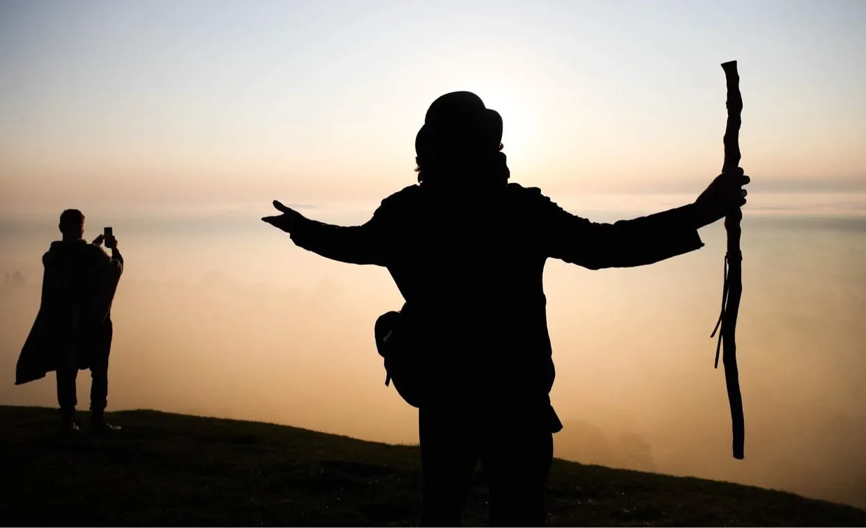 Tor Webster in silhouette on a hilltop in golden light, arms outstretched and staff raised above a sea of mist, with a visitor photographing the view in the distance.