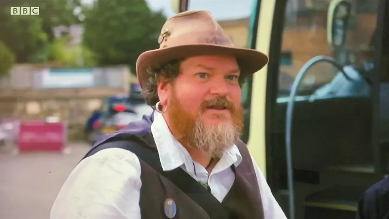 Tor Webster in a brown hat and waistcoat beside a bus, in a frame from his BBC One West appearance on The Great Staycation.