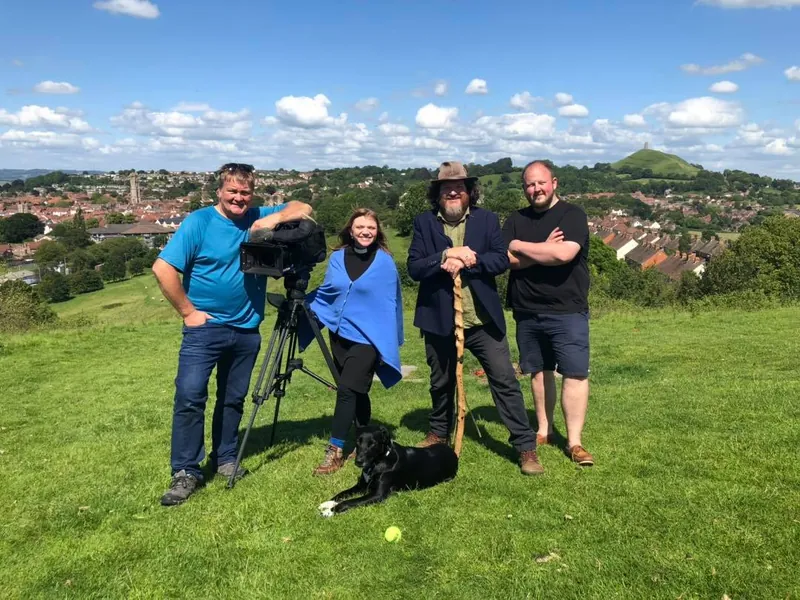 Tor Webster, in a hat and holding a staff, stands with a small film crew on a grassy hillside above Glastonbury while filming for the BBC. Glastonbury Tor rises on the skyline.